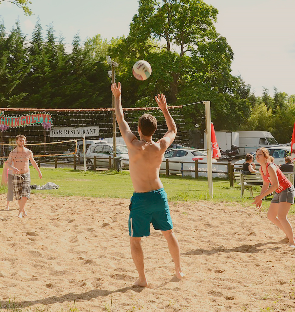 Sur cette image, on voit un homme de dos, en short, s'apprêtant à renvoyer un ballon par-dessus un filet de volley-ball. À droite, une femme en tee-shirt rouge semble prête à bondir, tandis qu'à gauche, un autre homme en short se tient juste en dessous de deux pancartes. Sur l'une est inscrit le mot 