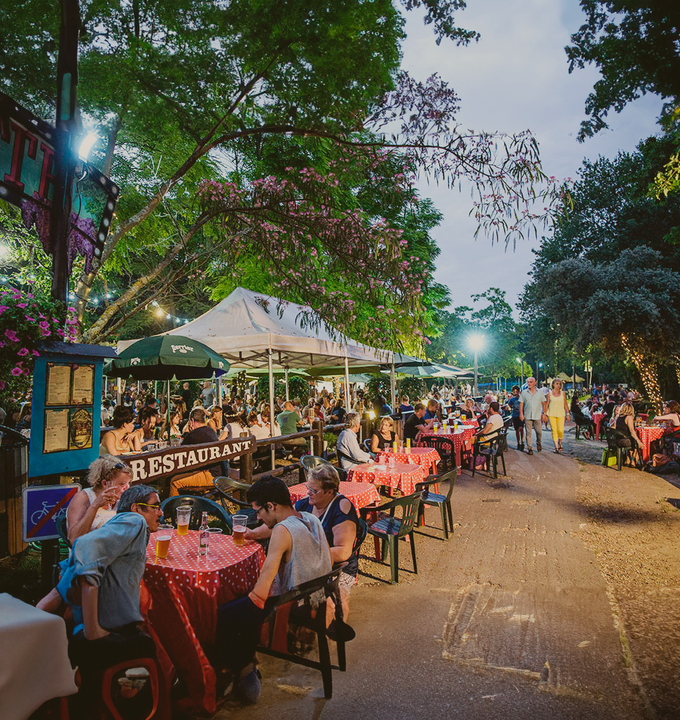 À gauche, au premier plan de l'image, quatre individus savourent un verre assis autour d'une table recouverte d'une nappe à carreaux rouges. Plus éloigné, au centre et légèrement en hauteur, un couple se promène le long de la Loire. L'atmosphère est rendue chaleureuse par la présence d'arbres, accentuant le charme de cette guinguette fréquentée par de nombreuses personnes installées aux tables, alors que les teintes du soir commencent à envelopper la scène.