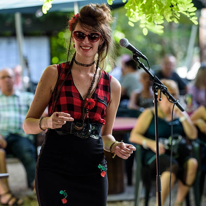 Vous découvrirez trois images, de gauche à droite, du même sujet. La première présente une chanteuse portant des lunettes de soleil en forme d'yeux de chat, un petit collier, et un débardeur à motif écossais rouge et noir. Elle est de face, souriante, avec un micro à sa droite. En arrière-plan, de manière légèrement floue, on distingue son public.