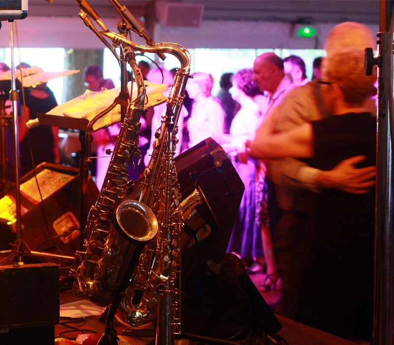 À gauche, au premier plan de l'image, deux saxophones dorés se distinguent. À droite, légèrement flous, un couple danse, et au fond, des personnes participent à ce qui semble être un thé dansant dans l'esprit d'un bal de guinguette.