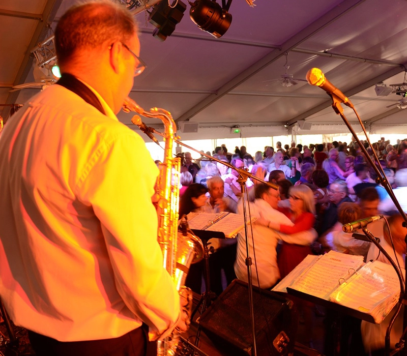 Au premier plan de l'image, un homme joue du saxophone. Dans le champ visuel de ce musicien, on aperçoit au moins 200 personnes dansant sous un chapiteau. Cette scène évoque l'ambiance d'un bal ou d'un thé dansant.