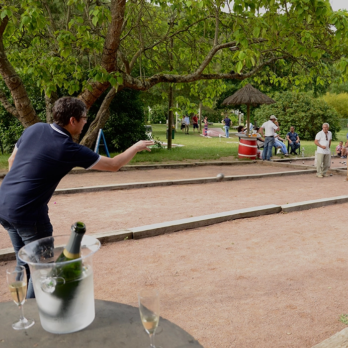 Dans cette scène, au premier plan, on remarque un seau de champagne contenant une bouteille, accompagné de deux coupes remplies, suggérant une atmosphère festive. Un peu plus en arrière-plan, en gros plan, se trouve un homme vêtu d'un polo bleu marine, vu de dos, en plein lancer d'une boule de pétanque. Il se positionne les pieds joints sur le terrain, concentré sur son jeu. Au loin, les autres joueurs observent, attendant patiemment leur tour pour entrer en action.